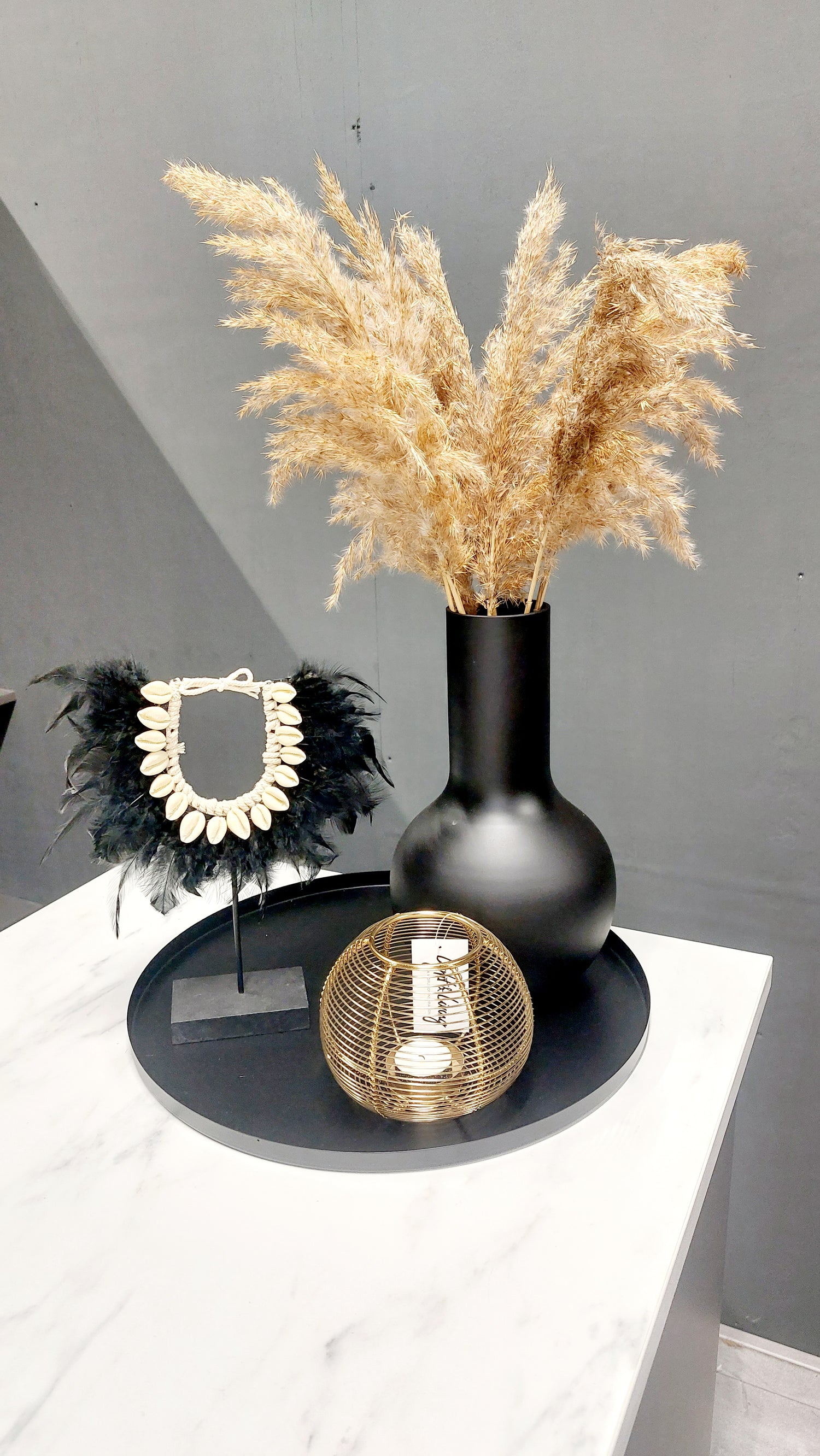Woonaccessoires set bestaande uit een zwart dienblad met daarop een mooi veren ornament een hoge vaas met pluimen en een gouden waxinelichthouder. 