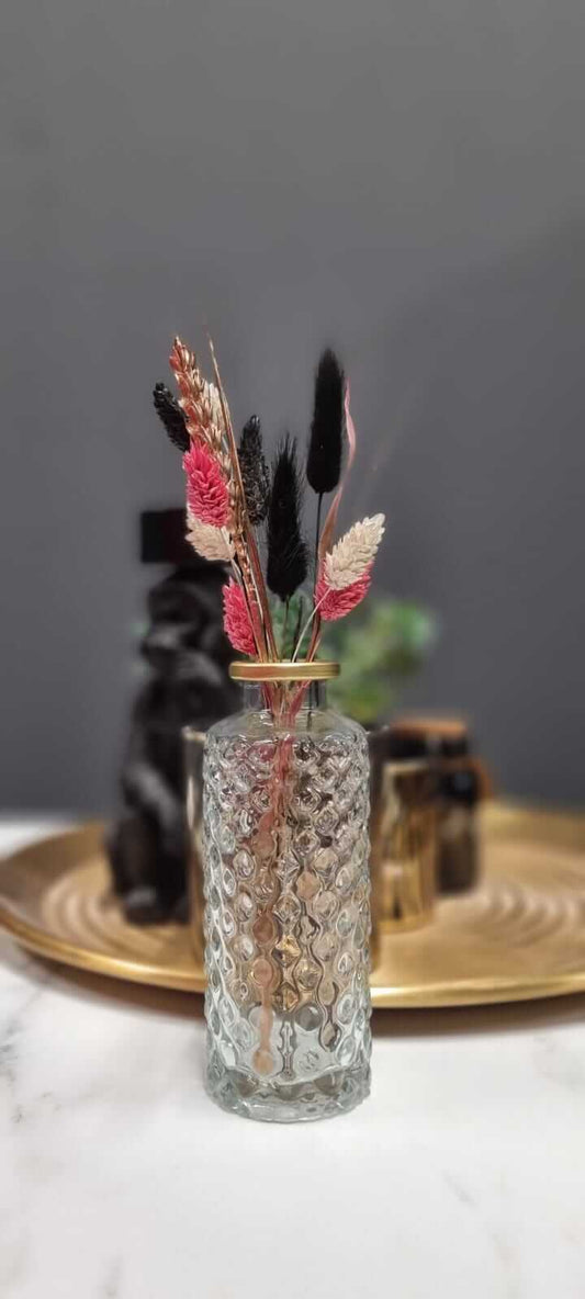 Elegant klein glazen vaasje met gouden rand, gevuld met een wisselende selectie droogbloemen. Perfect als tafeldecoratie of sfeervol cadeau.